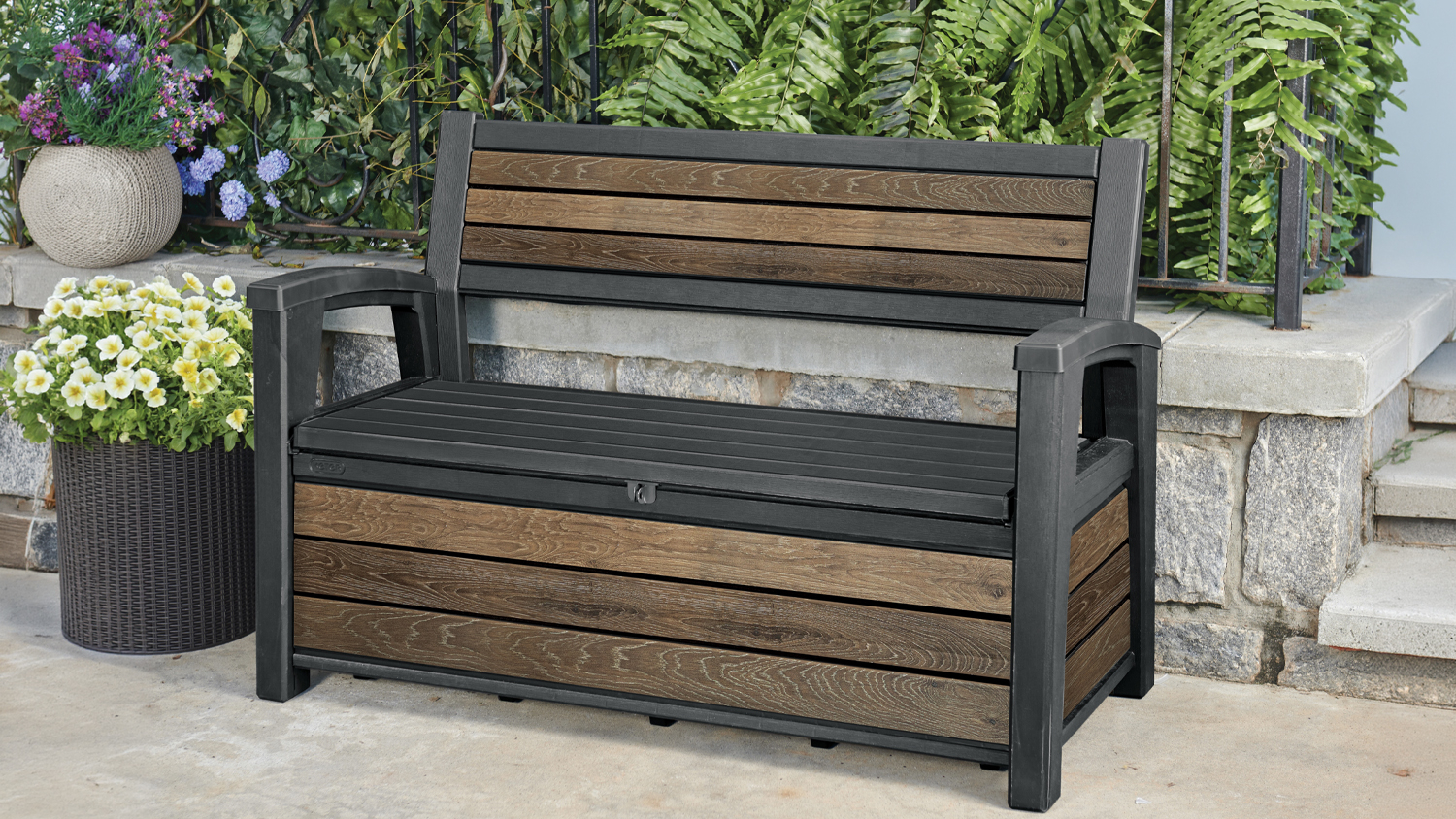 Keter Deco Small Storage Bench with wood-look panels and a black frame on a patio, surrounded by potted flowers and greenery