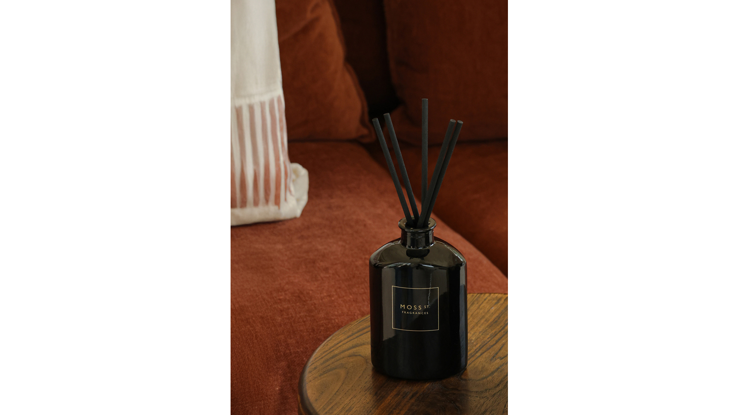 Close-up of a black Moss St. reed diffuser sitting on a wooden stool near a rust-coloured fabric sofa