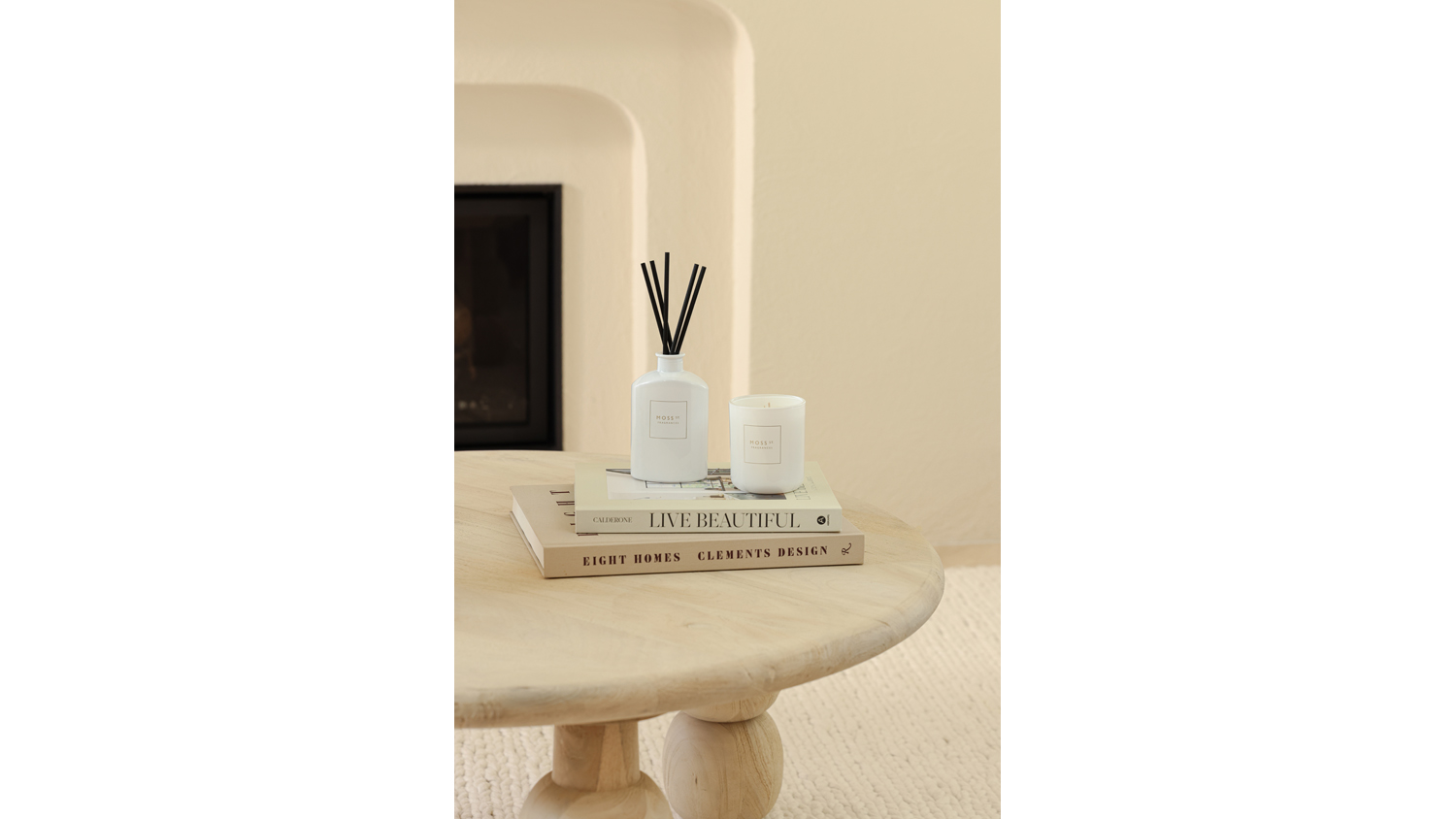 The white Moss St. mini reed diffuser and matching scented candle displayed on books atop a round wooden coffee table