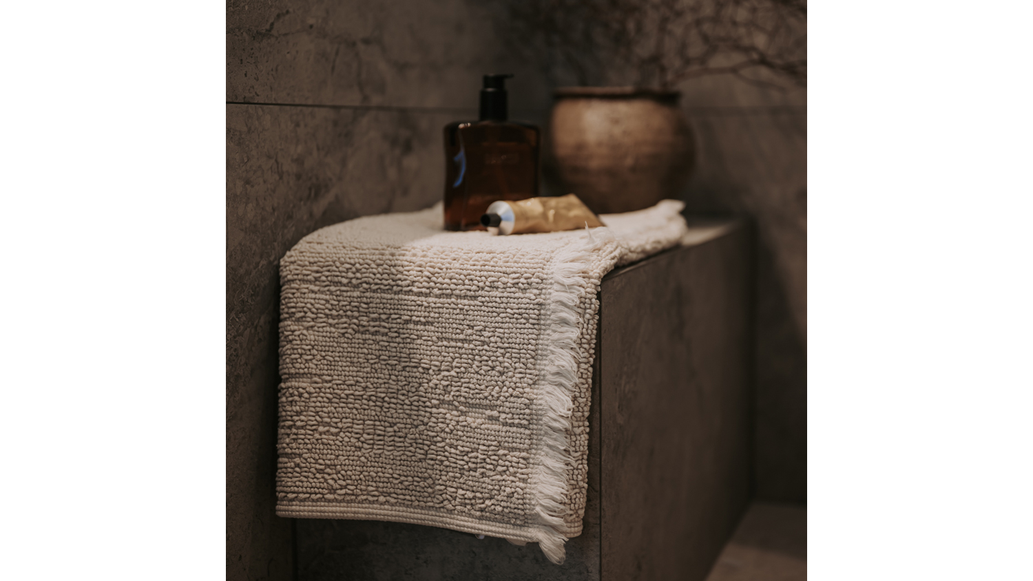 A linen-textured Baksana Lisbon Bath Mat is draped over a bathroom ledge beside toiletries and a decorative vase