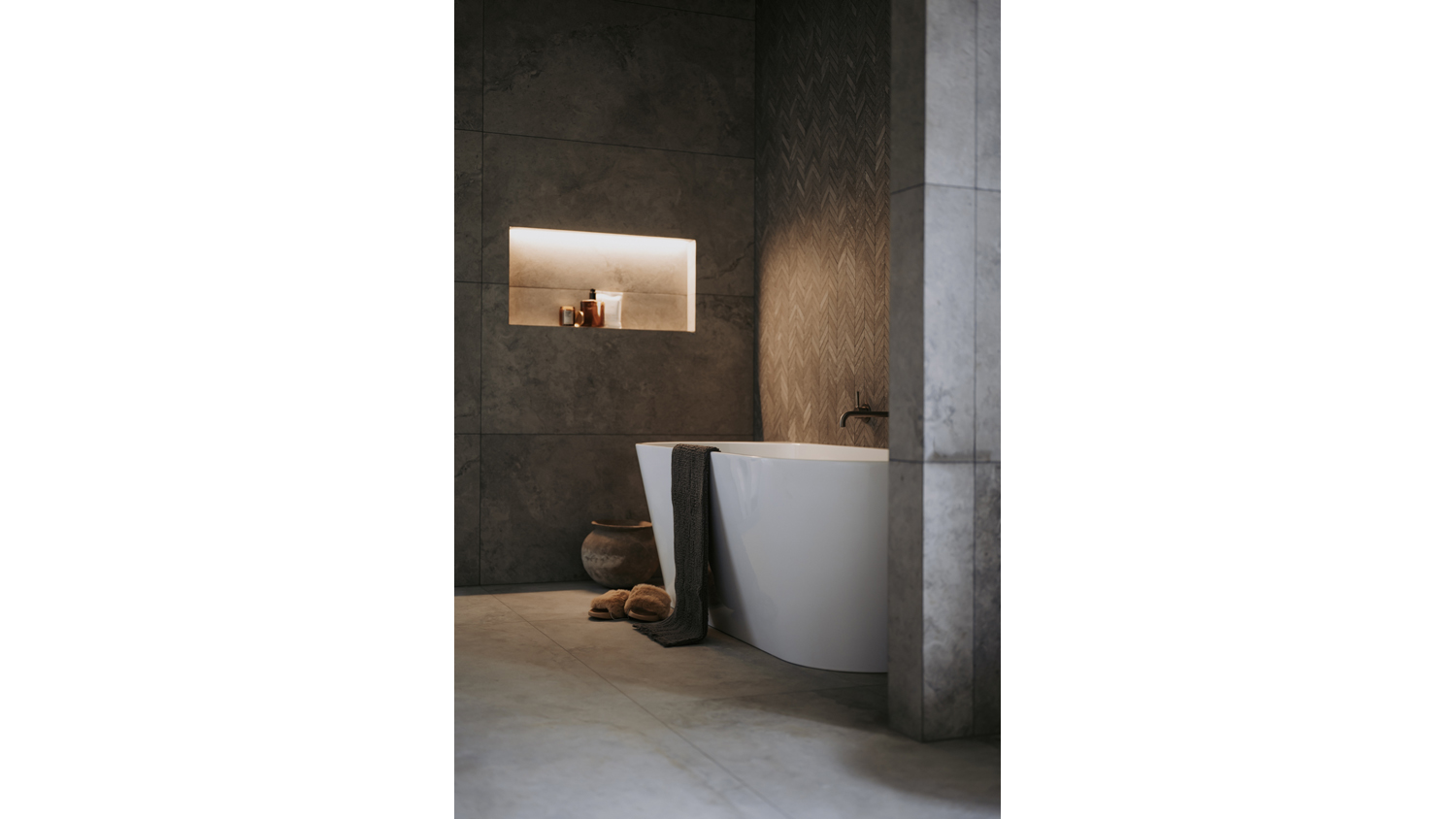 Modern bathroom with a freestanding white bathtub, dark stone tiles, and warm recessed shelf lighting