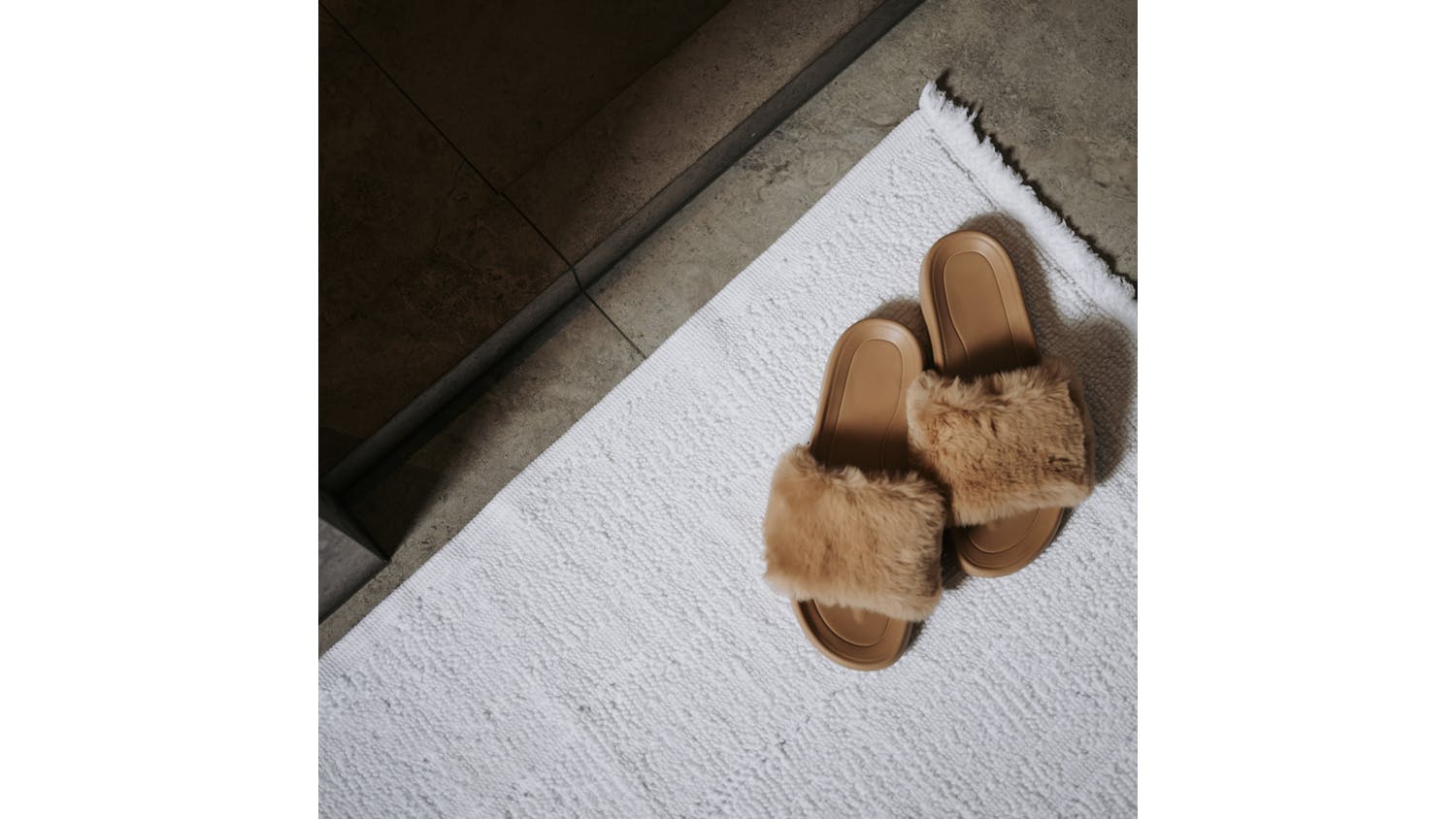 Pair of tan faux-fur slide slippers resting on a white Baksana Lisbon Bath Mat beside a tiled floor