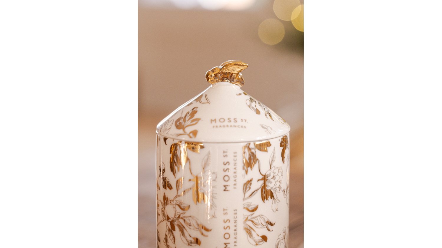 Close-up of gold bee-topped white ceramic candle with gold floral print, Moss St. branding, soft blurred background